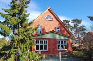 Uma casa de tijolos vermelhos com telhado verde e detalhes brancos, rodeada por árvores.