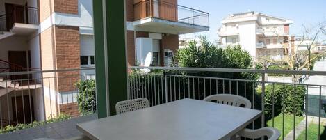 Uma varanda com mesa e cadeiras, um pilar verde e vista para edifícios vizinhos.