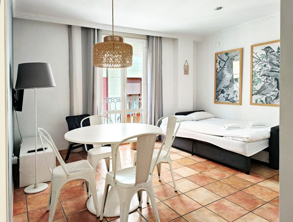 A hotel room with a round dining table, white chairs, a sofa bed, a hanging rattan light fixture, and two framed bird paintings on the wall.