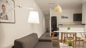 Moderni olohuone, jossa on harmaa sohva, ruokapöytä tuoleineen, keittiöalue kaappeineen ja katosta roikkuva valaisin.