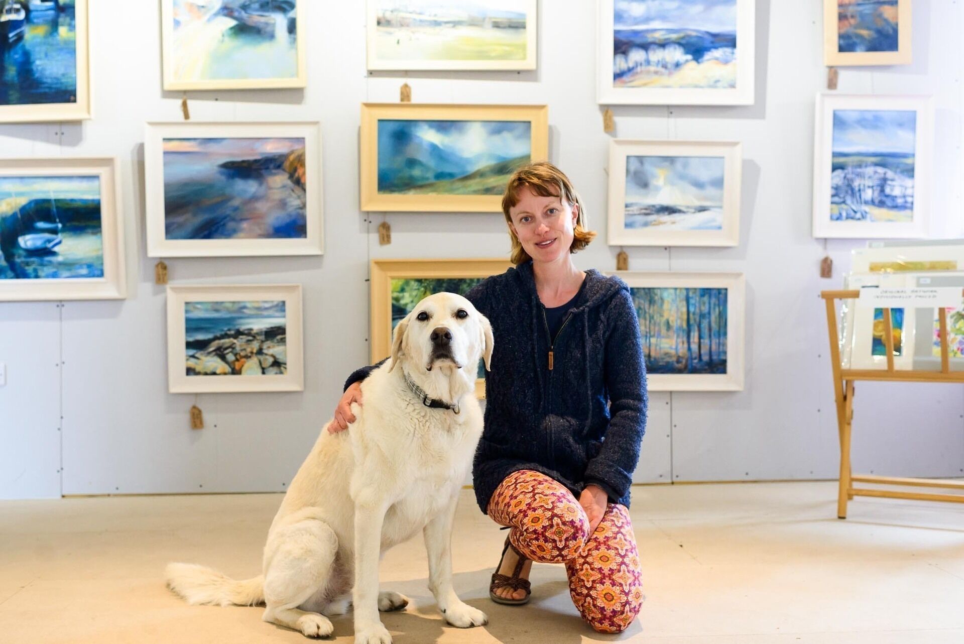Une personne est assise avec un chien dans une galerie d’art, les murs étant ornés de tableaux encadrés.