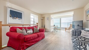 Amplia sala de estar con un sofá rojo, un sofá estampado y un televisor. Hay un comedor con mesa y sillas, y un gran ventanal con vistas.