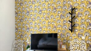 Ein Zimmer mit einem Fernseher auf einem weißen Schreibtisch, zwei weißen Stühlen und gelbem Blumenwallpaper.