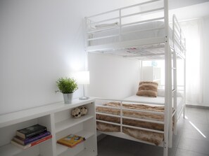 A bunk bed room with a white wall, a small shelf with books, a potted plant, and a lamp.