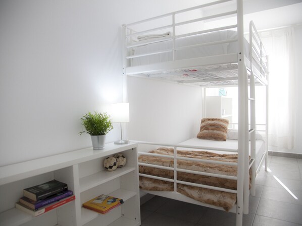 A bunk bed room with a white wall, a small shelf with books, a potted plant, and a lamp.