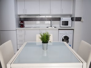 A modern kitchen with white cabinets, a microwave, a washing machine, and a glass table with a potted plant. - Flats Friends Tetuan 2 bedrooms (Valencia)