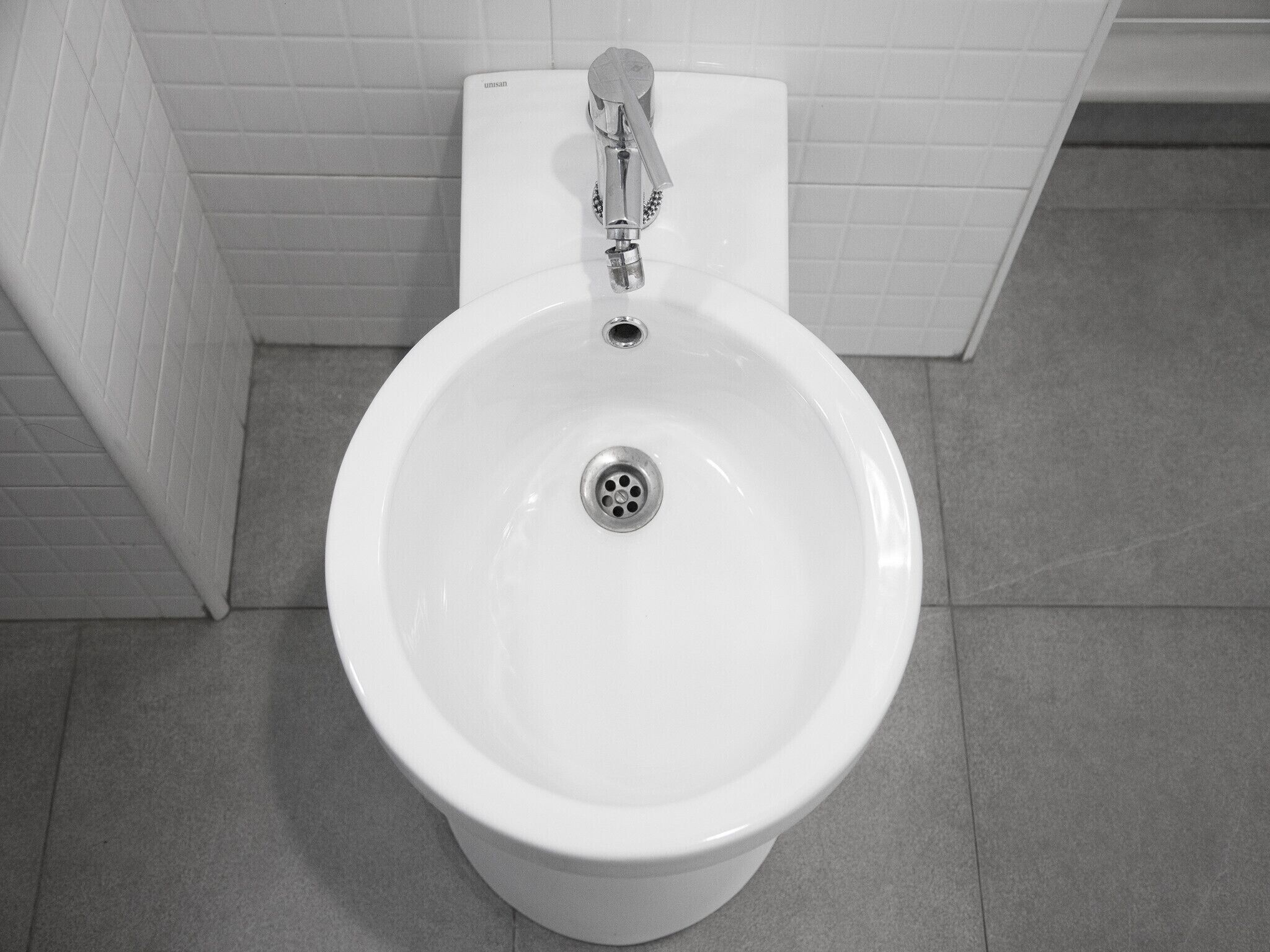 A white bidet with a chrome faucet and a drain in a bathroom with white tiled walls and floor.