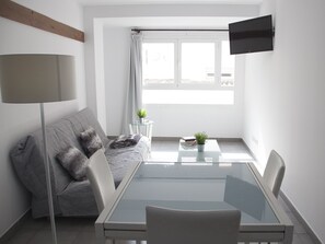 A modern, minimalist living room with a grey sofa, a glass coffee table, and a flat-screen TV mounted on the wall.