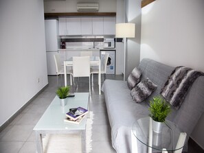 A modern living room with a grey sofa, a glass coffee table, and a dining area with white chairs and a table.