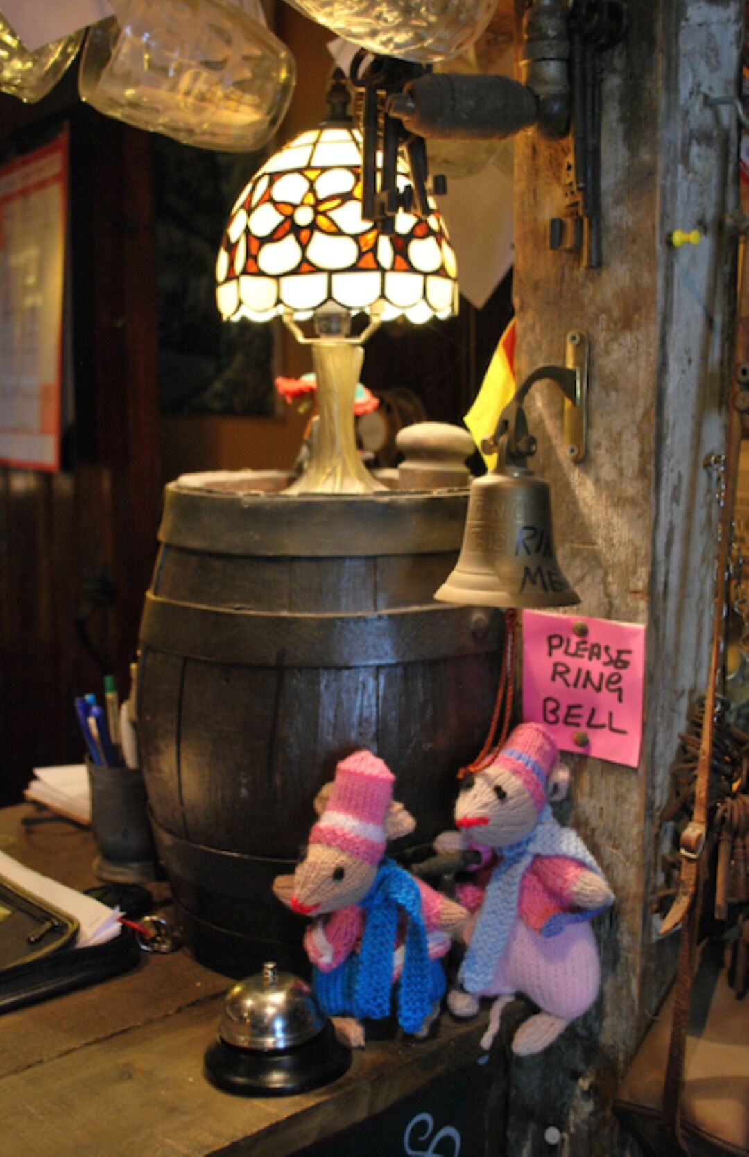 Un interior rústico con un barril de metal grande, una lámpara de vitral y una campana. Hay dos figuras de animales tejidos y un cartel que dice 'Por favor, toque la campana'.