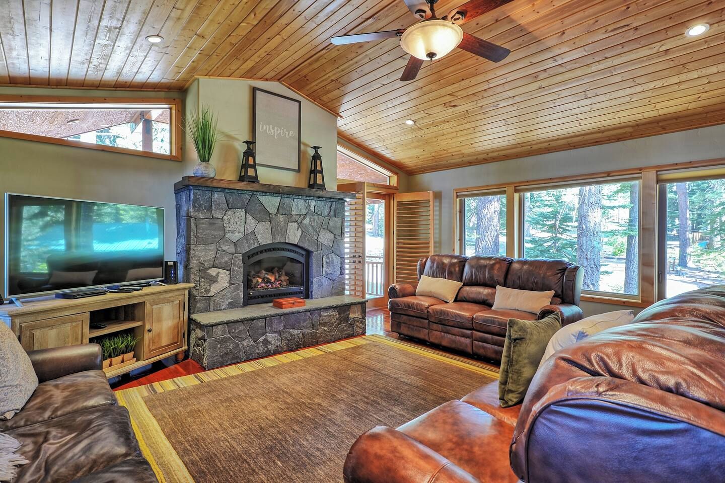 A cozy living room with a stone fireplace, leather sofas, a flat-screen TV, and large windows offering a view of the outdoors.