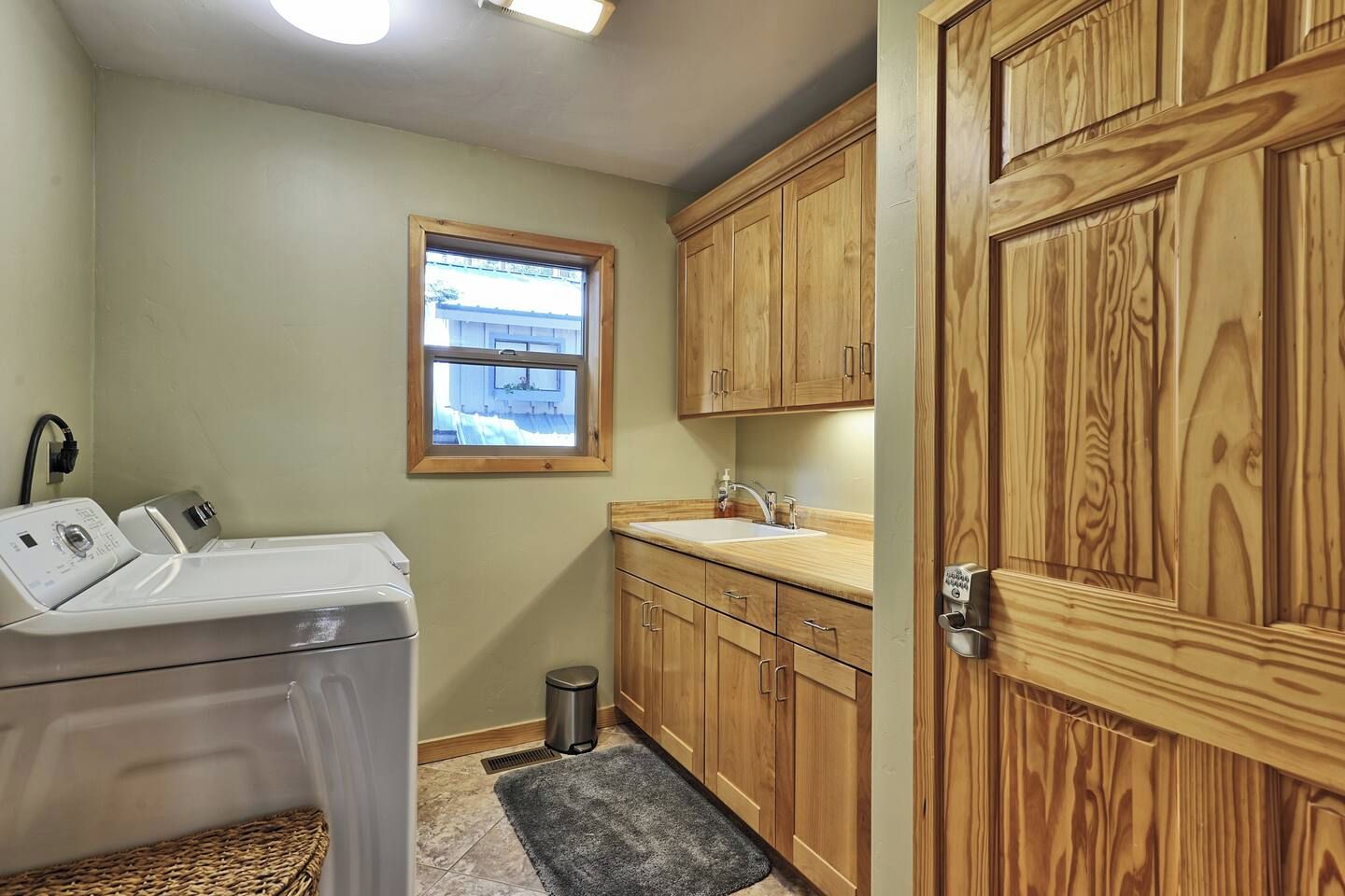 A laundry room with a washing machine, a sink, wooden cabinets, a window, and a trash can.