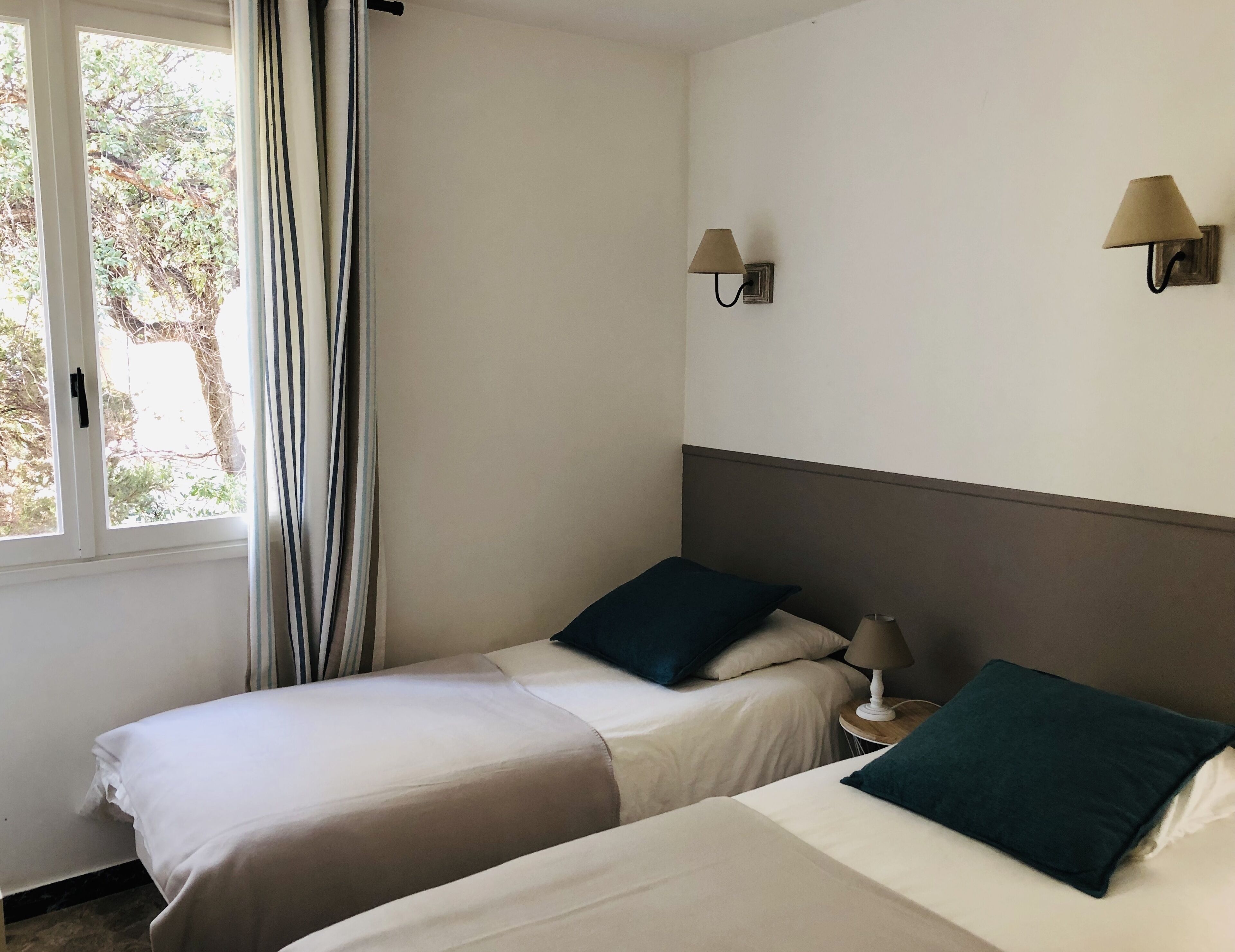 A hotel room with two beds, a window with curtains, and two wall-mounted lamps.