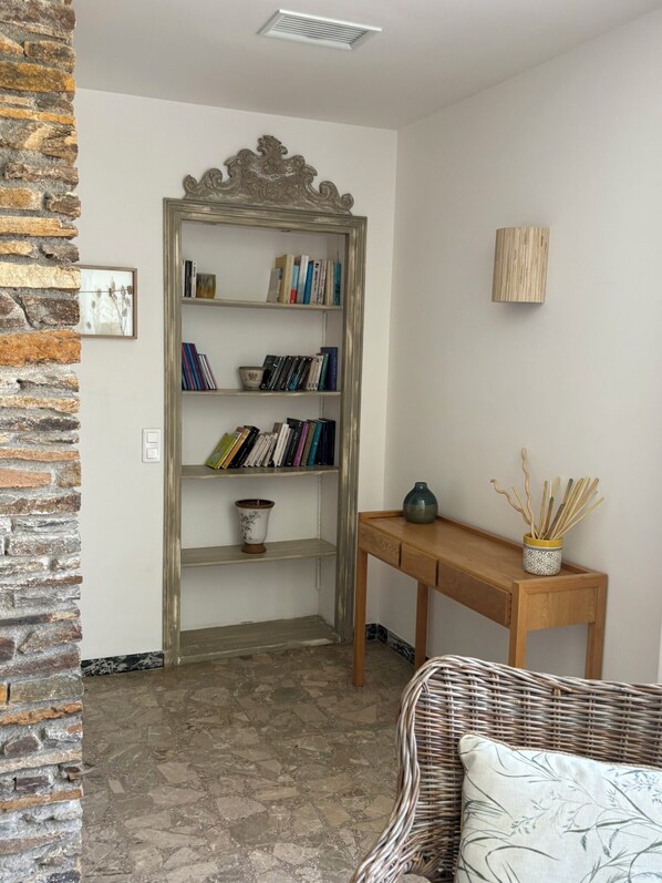 Una habitación con pared de piedra, una estantería, una mesa de madera y una silla de mimbre.