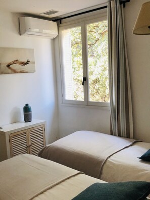 A bedroom with two beds, a window with curtains, a wall-mounted air conditioner, a bedside table with a vase, and a painting. - Tropics Villa - Sea view - Heated pool 28 ° C (Cavalaire-sur-Mer)