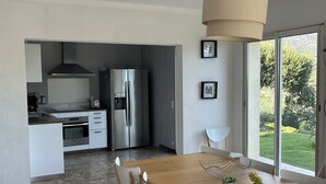 Une cuisine moderne équipée d’un réfrigérateur en acier inoxydable, d’un four encastré et d’une machine à café. On y trouve également un coin repas avec une table en bois et des chaises blanches.