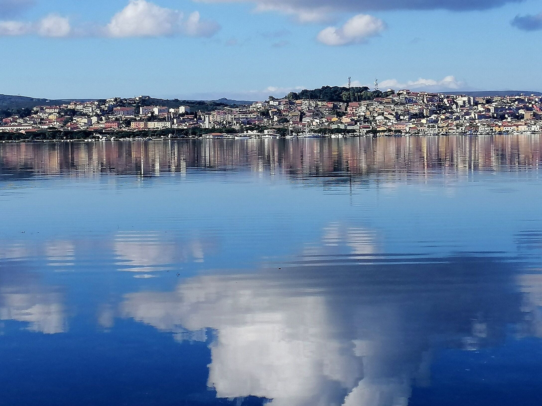Une étendue d’eau calme qui reflète une agglomération densément peuplée, avec des bâtiments et une colline à l’horizon.
