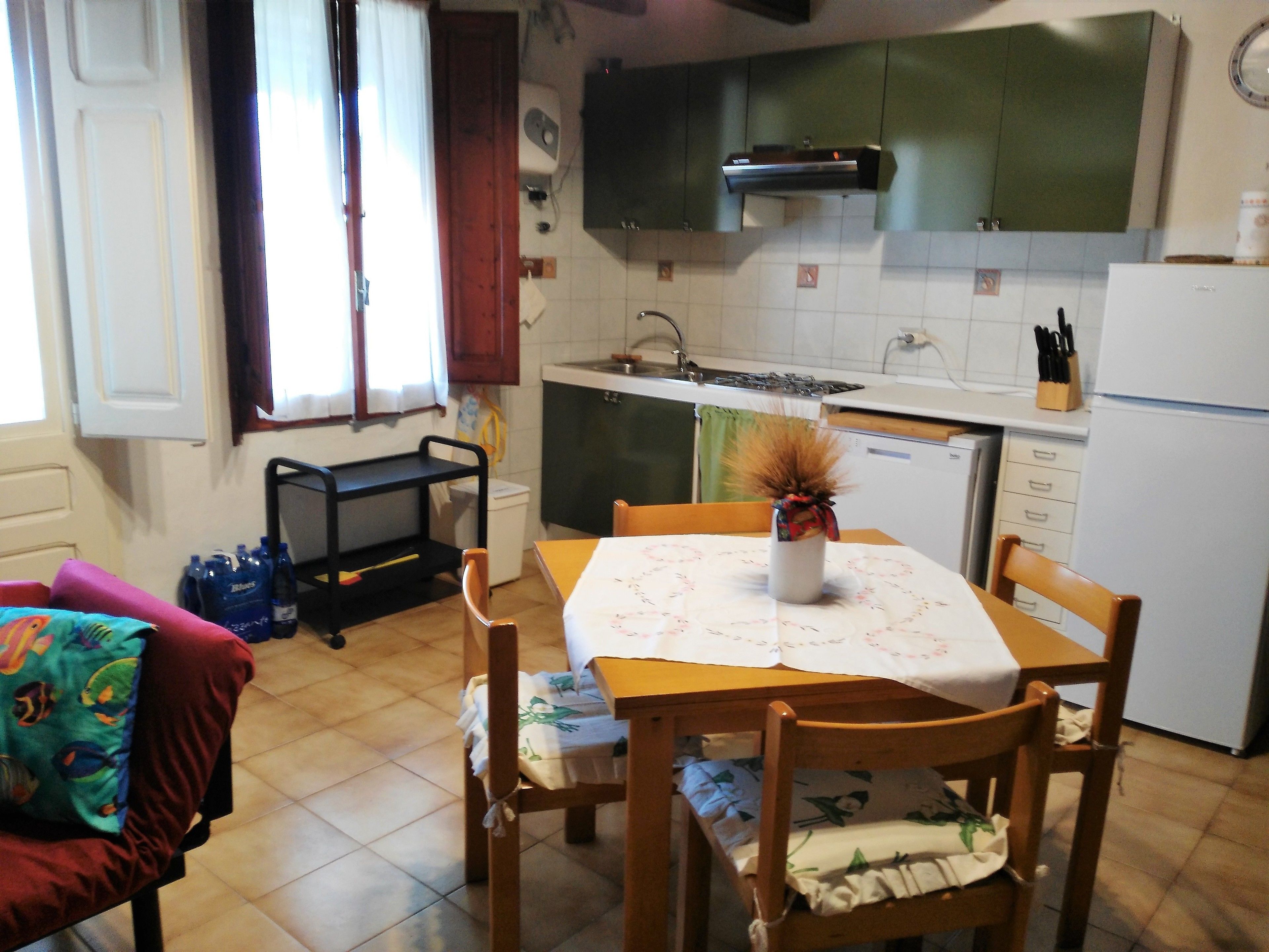 Una cocina con una mesa de comedor, sillas, un florero con flores secas, un refrigerador y una ventana con cortinas.