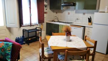 Una cocina con una mesa de comedor, sillas, un florero con flores secas, un refrigerador y una ventana con cortinas.