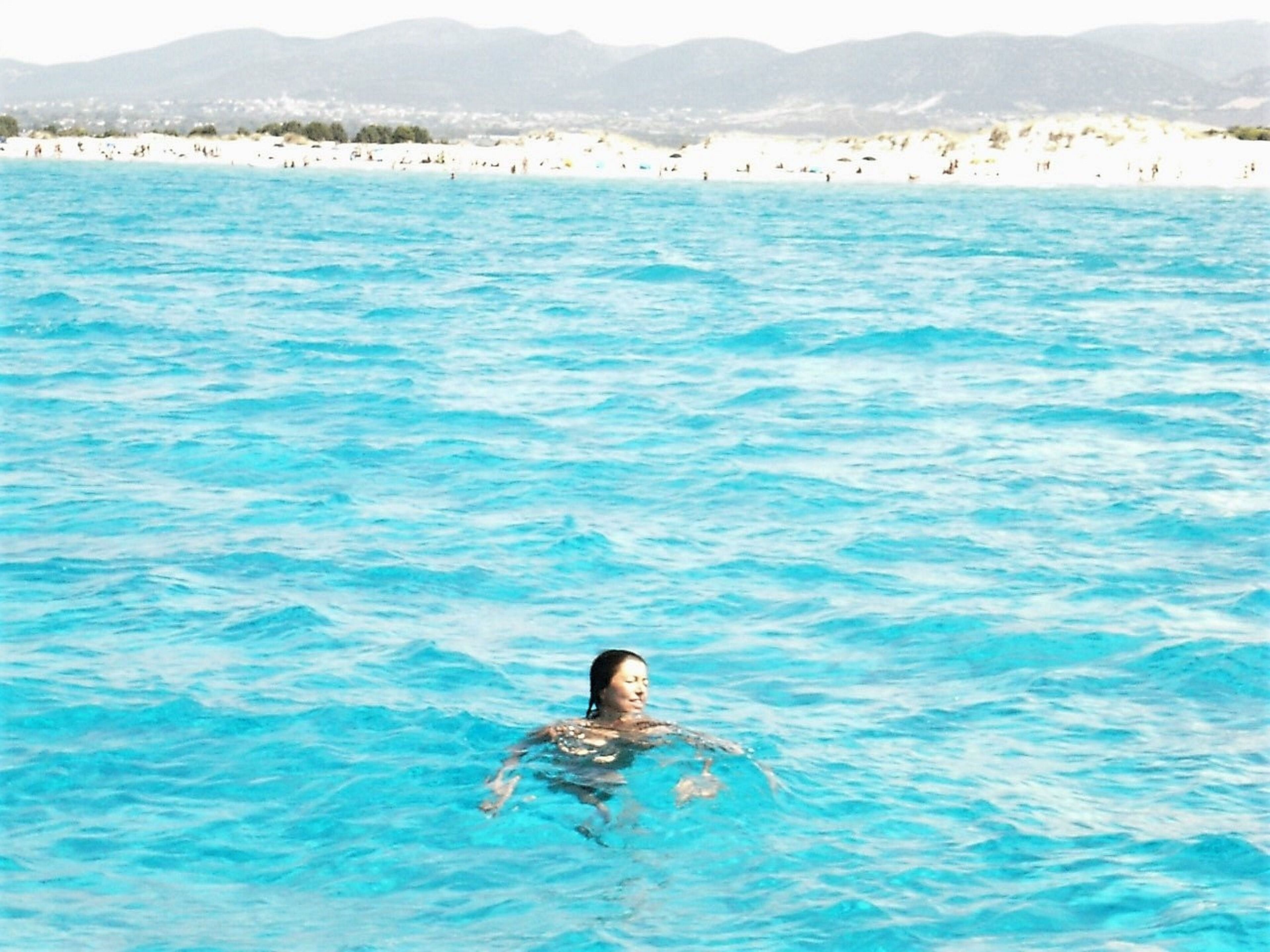 Iemand zwemt in helderblauw water, met een strand en bergen op de achtergrond.