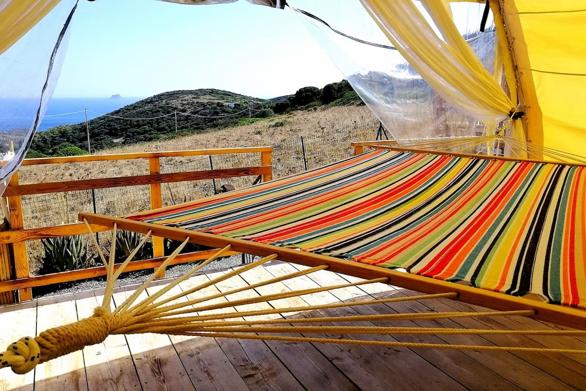 Ein Hängestuhl mit bunt gestreifter Stoffbespannung, auf einer Holzterrasse platziert, mit Blick auf eine hügelige Landschaft und das Meer.