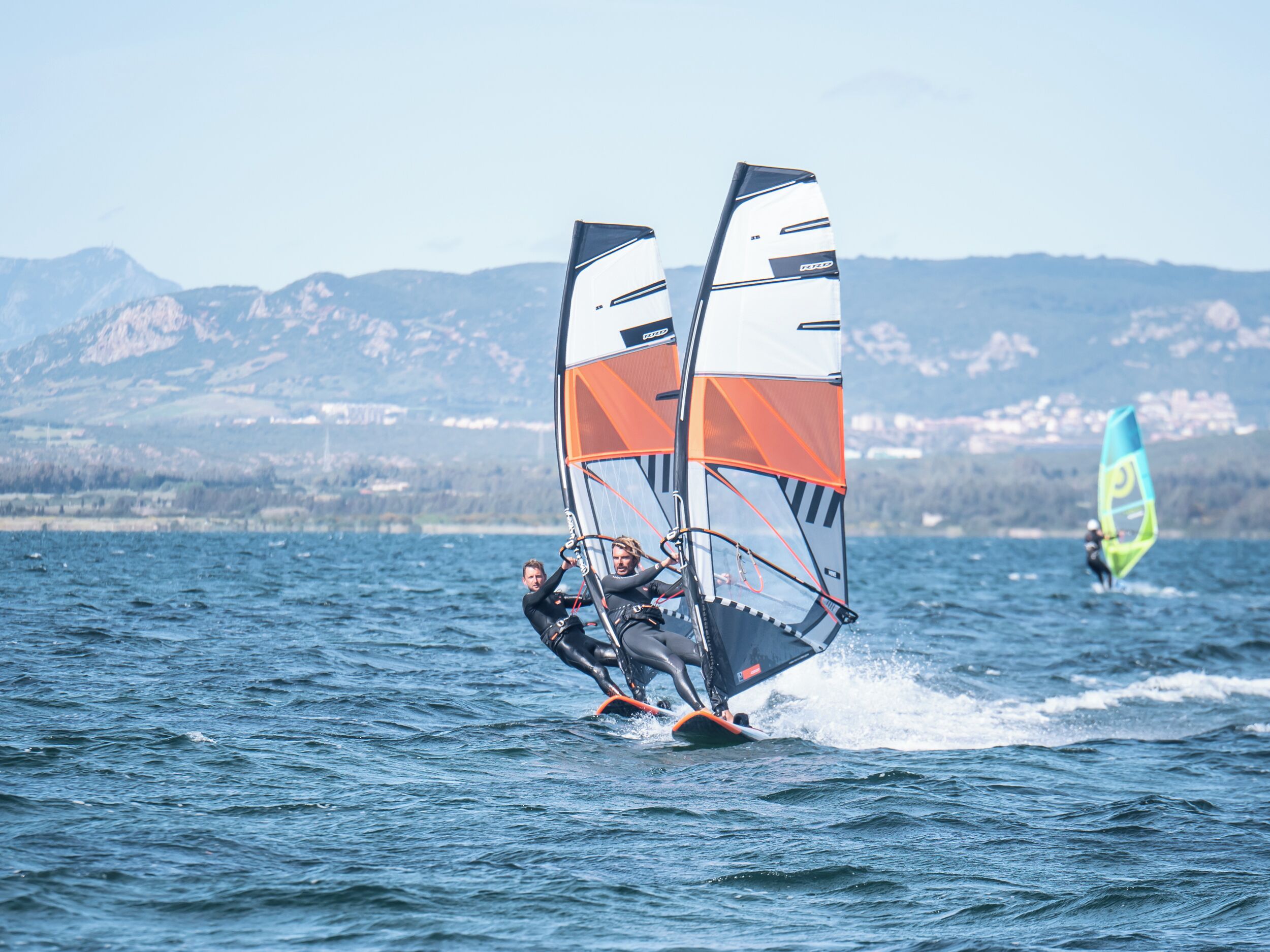 Una persona practica windsurf en un lago con montañas al fondo.