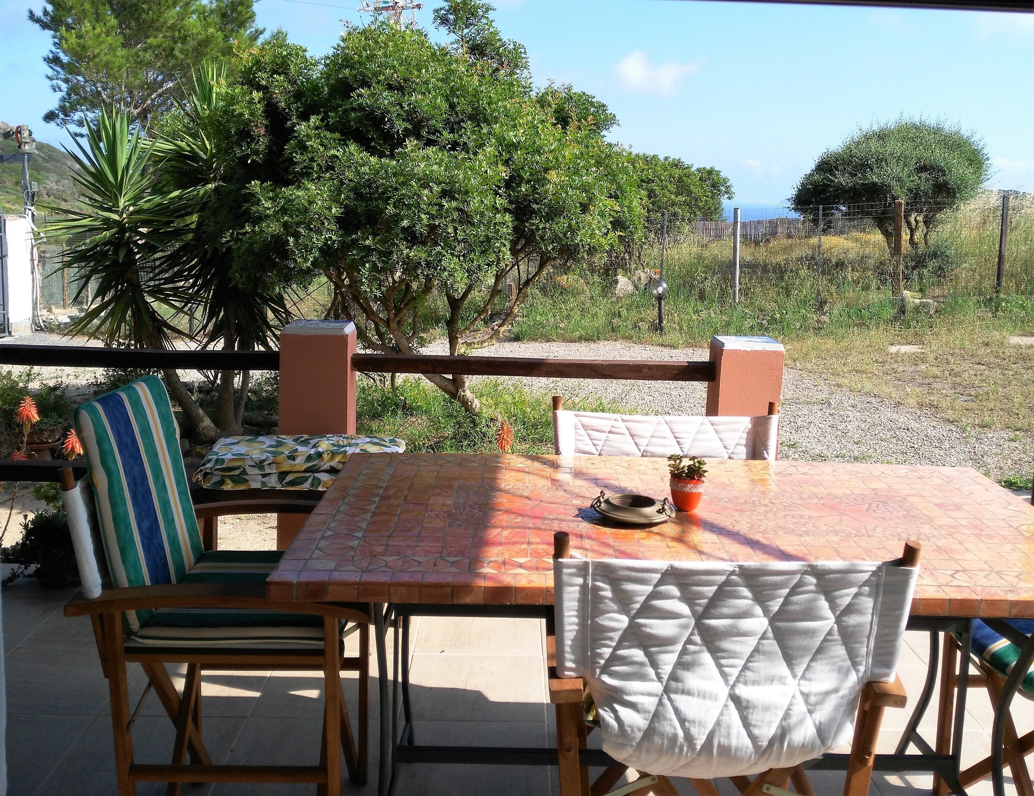 An outdoor patio with a tiled table and two chairs, surrounded by greenery and a view of the landscape.