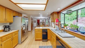 Una cocina moderna con armarios de madera, electrodomésticos de acero inoxidable, lavavajillas y un amplio ventanal con vistas a árboles.