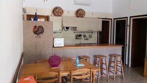 Una cucina con un tavolo di legno, sedie e uno sgabello da bar. Si nota una ventola a soffitto, un condizionatore d'aria e un gallo decorativo appeso al muro.