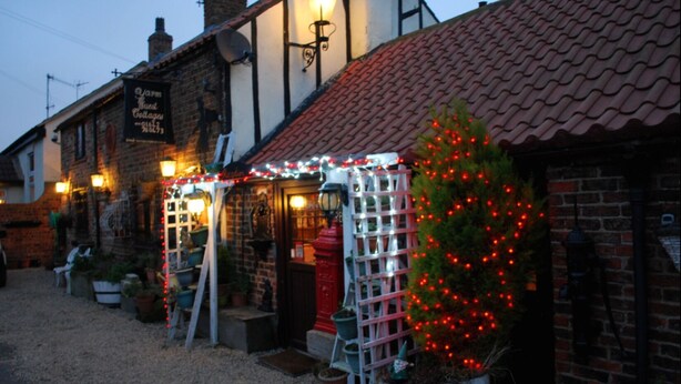 Un bâtiment traditionnel en briques, avec un toit recouvert de tuiles, une enseigne et un sapin de Noël décoré de guirlandes lumineuses rouges.