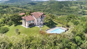 Une grande maison avec une piscine, entourée d’une végétation luxuriante et avec des collines en arrière-plan.
