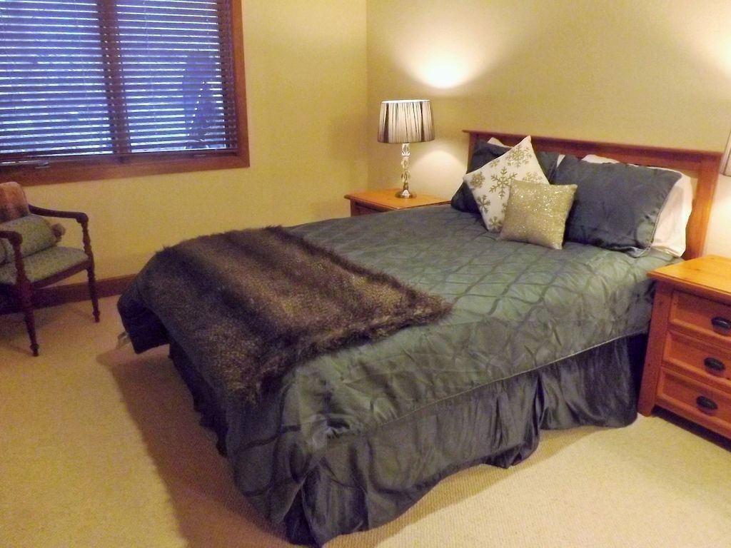 A bedroom with a bed, a nightstand, a lamp, a chair, and a window with blinds.