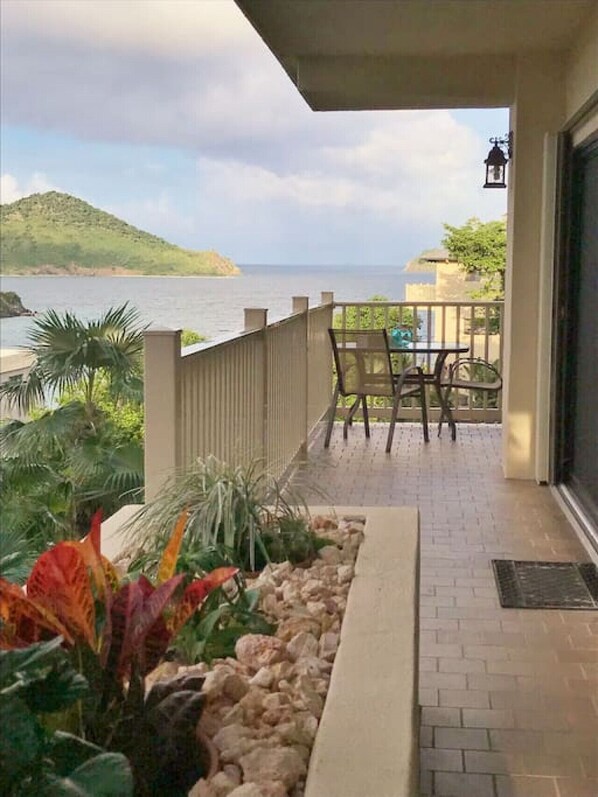 A balcony with a view of the sea, a table with chairs, and a planter with tropical plants.