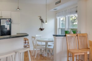 Una cocina con armarios blancos, un refrigerador, una mesa de comedor con sillas blancas y una ventana con vista a árboles.