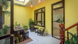 Um quarto amarelo e claro com uma mesa de jantar de madeira, cadeiras e plantas em vasos. Há uma janela grande e uma escada de madeira com corrimão.