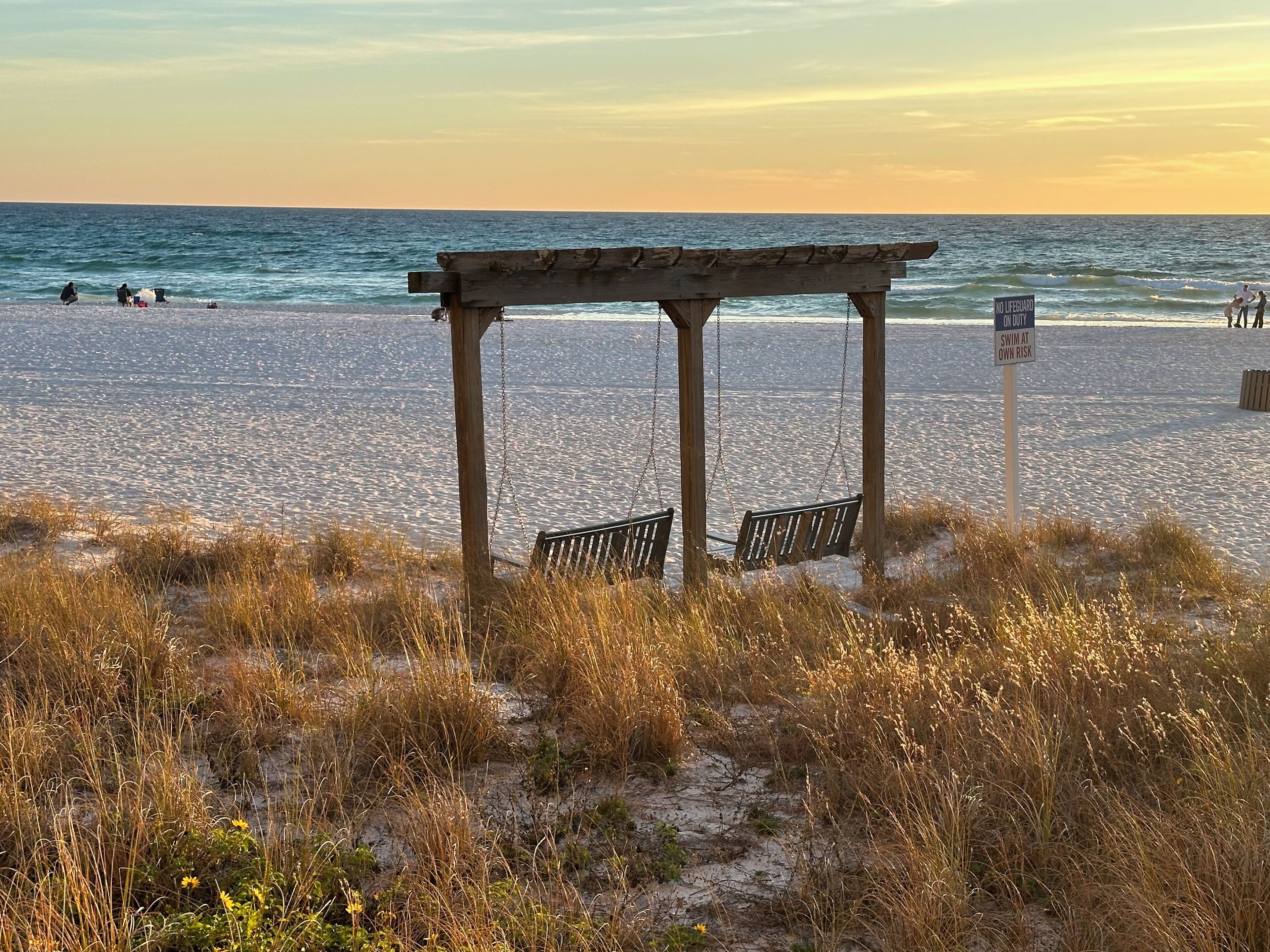 Une plage avec une structure en bois, un panneau indiquant l’absence de sauveteur, et des personnes dans l’eau.