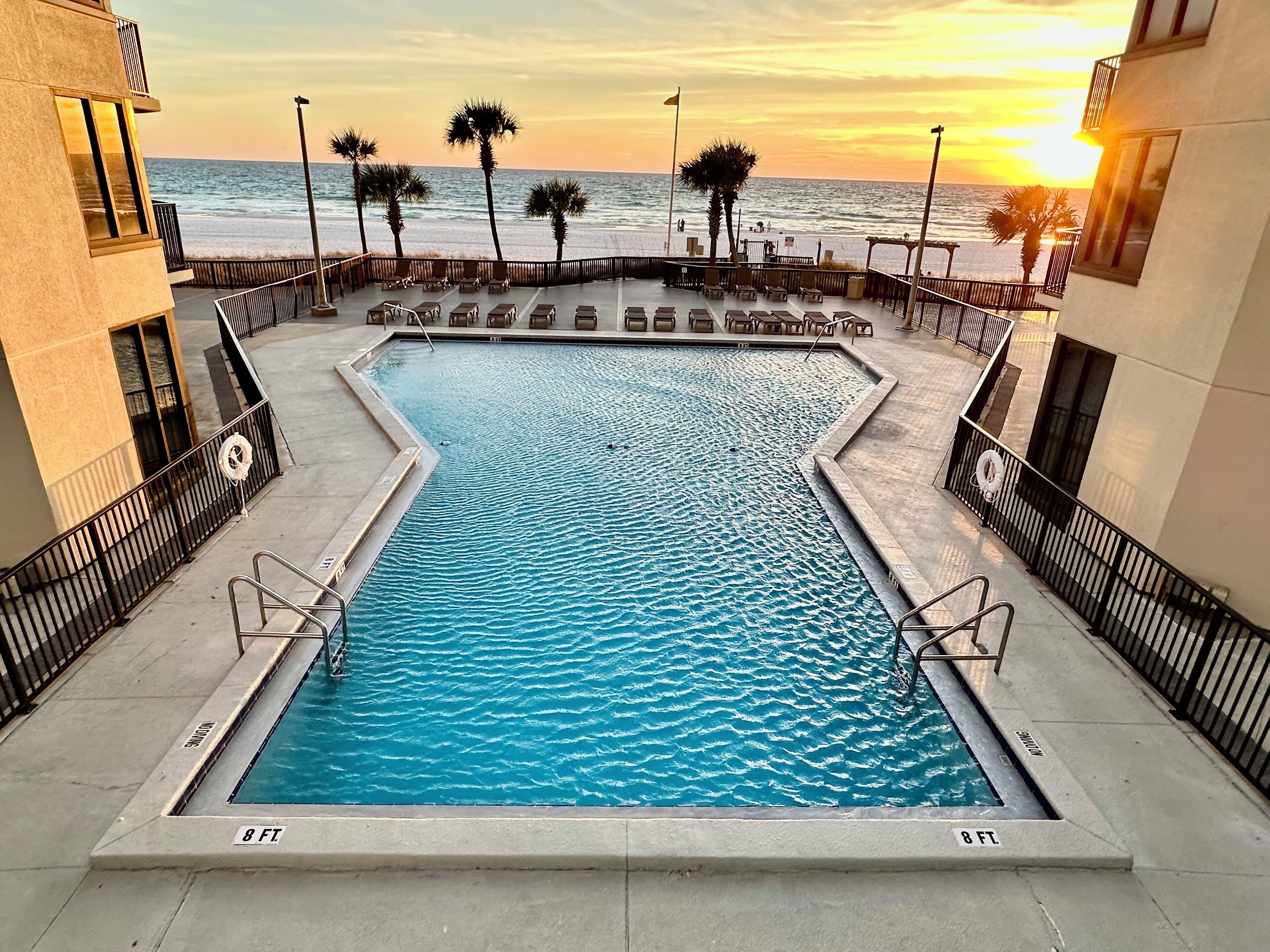 Une piscine rectangulaire d’une profondeur de 2,4 mètres, entourée de chaises longues et de palmiers, offrant une vue sur l’océan et le coucher de soleil.