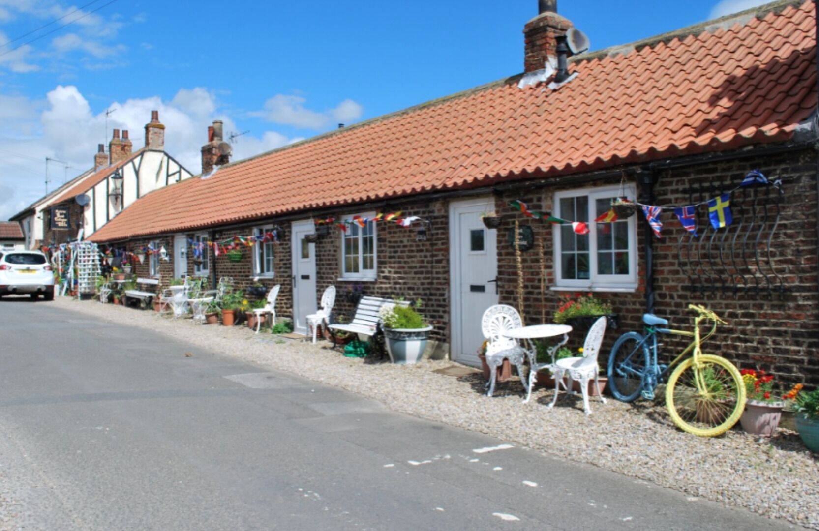 A row of traditional brick houses with red-tiled roofs, white doors, and windows. There are outdoor seating areas with white metal chairs and tables, and a bicycle parked in front of one house.