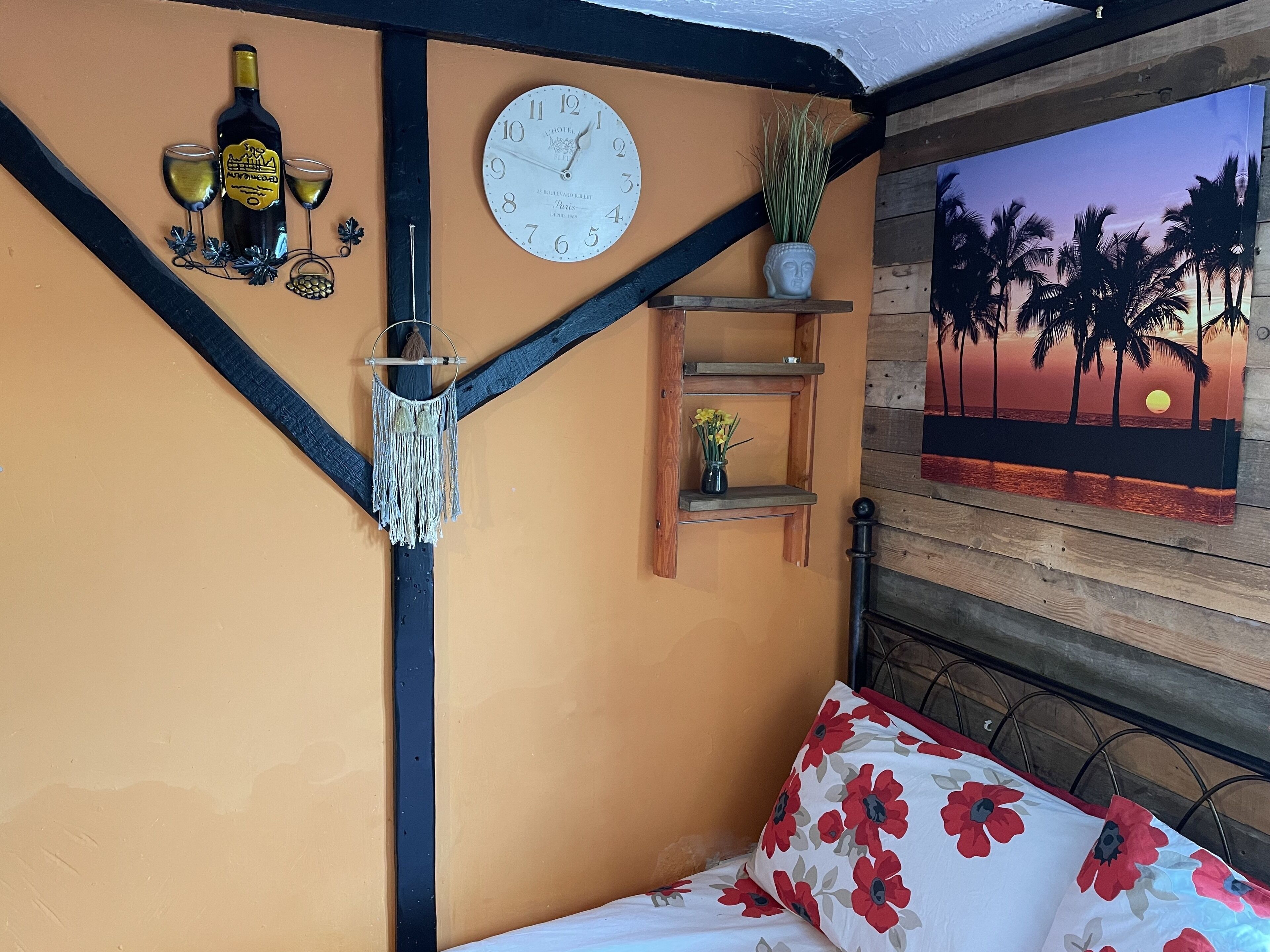 Une pièce comprenant un lit, une horloge murale, une bouteille de vin, des verres, une décoration en macramé, une petite étagère avec une plante et un tableau représentant un coucher de soleil.