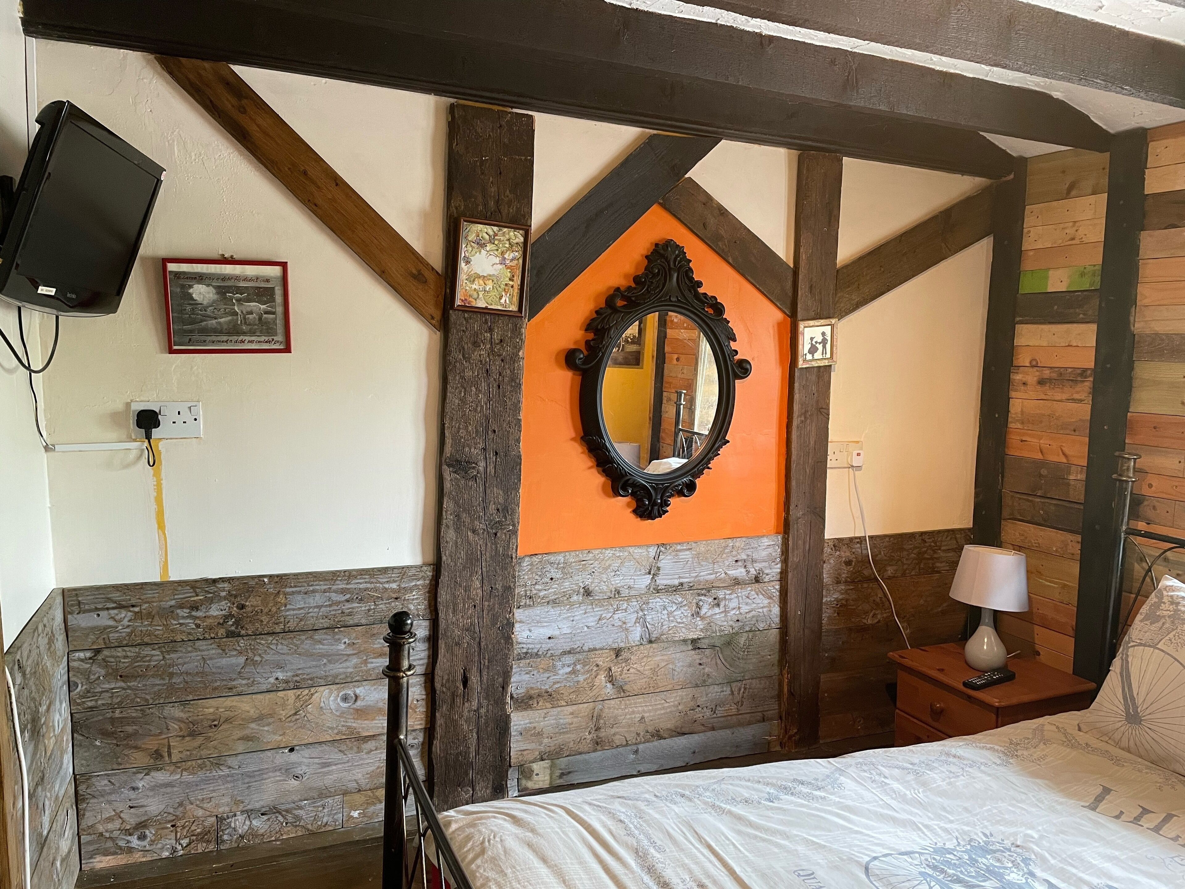 Une chambre à coucher avec une tête de lit en bois, un lit recouvert d’une couette blanche, une table de chevet avec une lampe, un téléviseur fixé au mur et un tableau encadré.