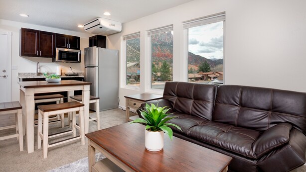 A modern kitchen with dark cabinets, a stainless steel refrigerator, and a microwave. There is a dining area with wooden chairs and a table, and a brown leather sofa in the living area. A potted plant is on the coffee table. - Apartment #3 GREAT LOCATION Zion National Park entrance as close as you can get! (Springdale)
