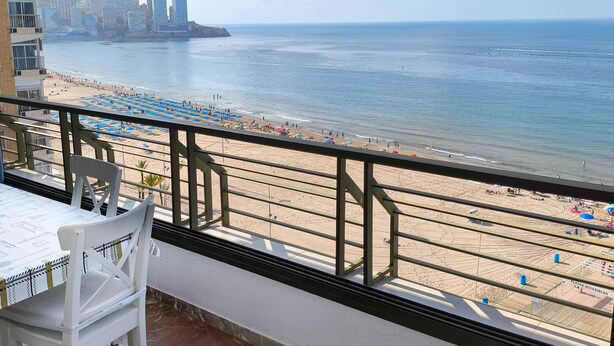 Un balcón con vistas a la playa, un área de comedor con mesa y sillas, y un paisaje urbano de fondo.