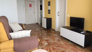 Una sala de estar con un sofá, un televisor sobre un mueble bajo, una mesita y una puerta que da a otra habitación.