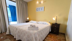 Habitación de hotel con cama doble, dos lámparas de noche, una mesita auxiliar pequeña, un cuadro en la pared y vistas a través de una ventana con cortinas.