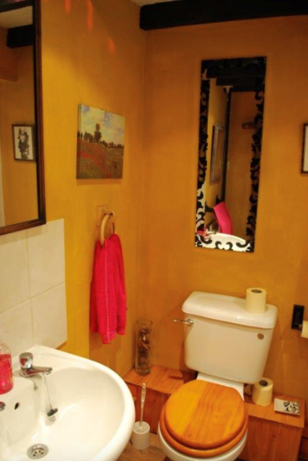 Une salle de bain avec un lavabo blanc, un abattant de toilettes en bois, un miroir à cadre noir et un tableau accroché au mur.