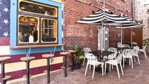 An outdoor seating area with tables and chairs, a food service window, and a brick wall. - 5th-Floor Condo | Balcony + Parking | Heart of AVL (Asheville)