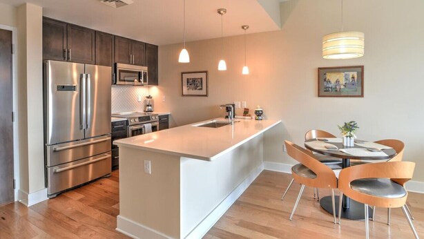 A modern kitchen with stainless steel appliances, a central island, and a dining area with wooden chairs. - 5th-Floor Condo | Balcony + Parking | Heart of AVL (Asheville)