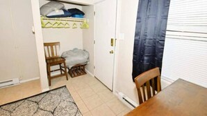 A room with a dining table, a chair, a small table, a bench, a closet with hanging clothes, and a door.
