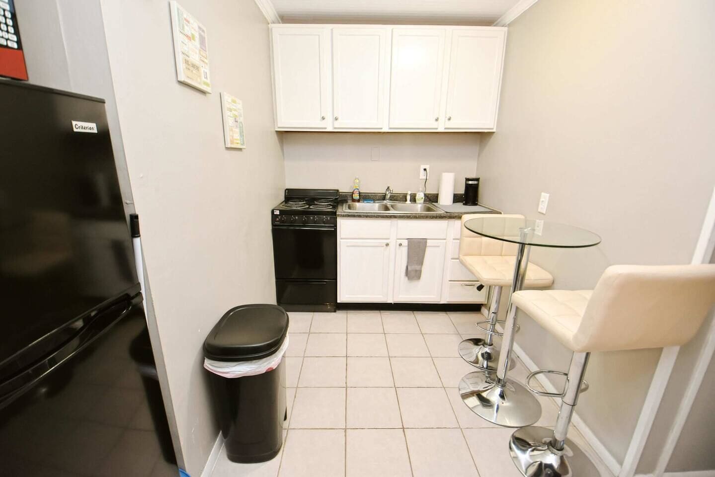 A compact kitchen with white cabinets, a black stove, a trash bin, and a glass table with two chairs.