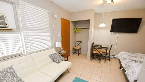 Ein Zimmer mit weißem Sofa, einem Bett, einem Esstisch mit Stühlen, einem an der Wand befestigten Fernseher und einem Fenster mit Jalousien.